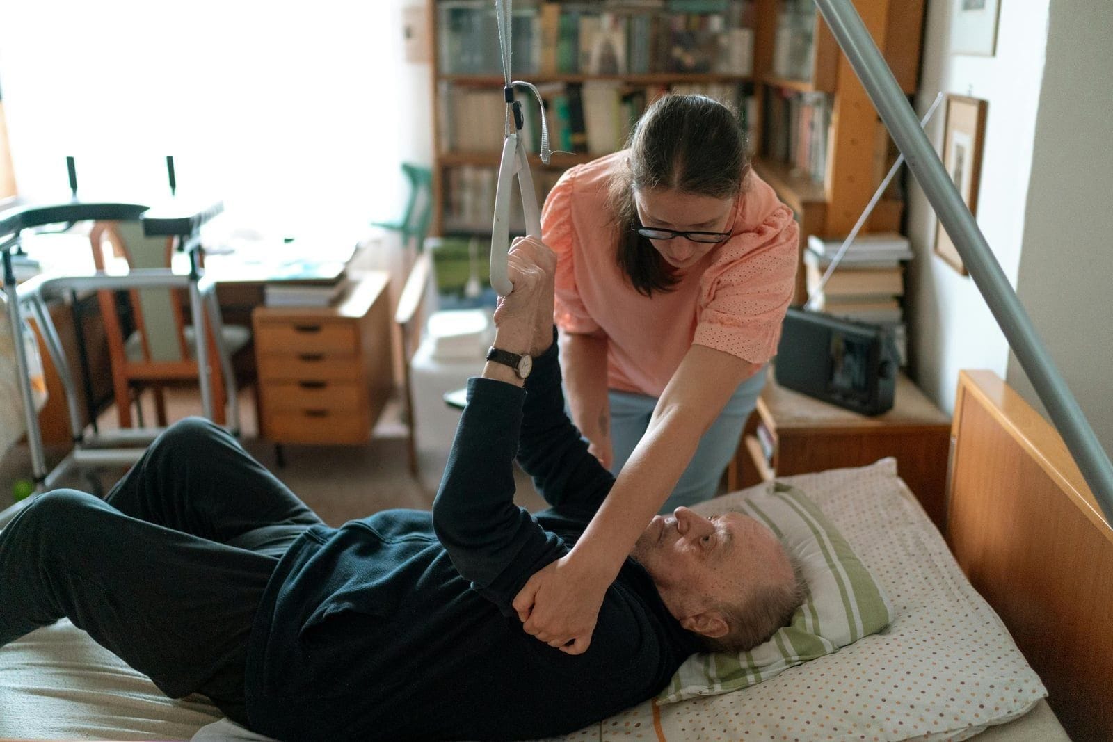 A home caregiver in a soft pink blouse leans over a hospital-style bed to help an elderly man lift his arms toward an overhead trapeze handle, his thin body resting on a pillow patterned with green stripes, the room a familiar mix of bookshelves and a folded walker that signals home care has settled into the daily rhythm of advanced Parkinson's