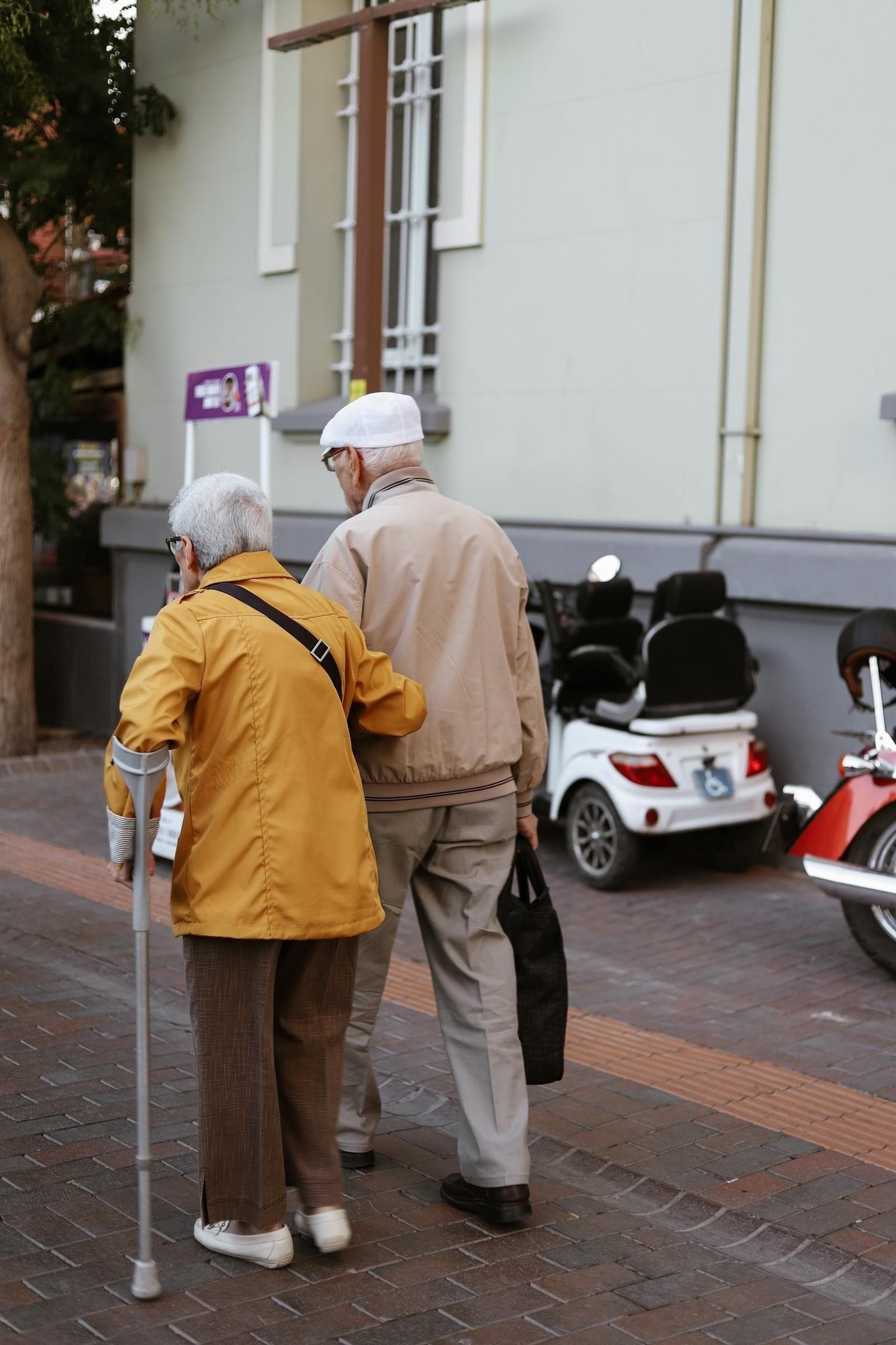 A senior couple walks slowly down a sunlit street, the husband leaning gently on a wooden cane, his wife's hand resting lightly at the small of his back, the unhurried pace of two people who have learned that the way through Parkinson's is together