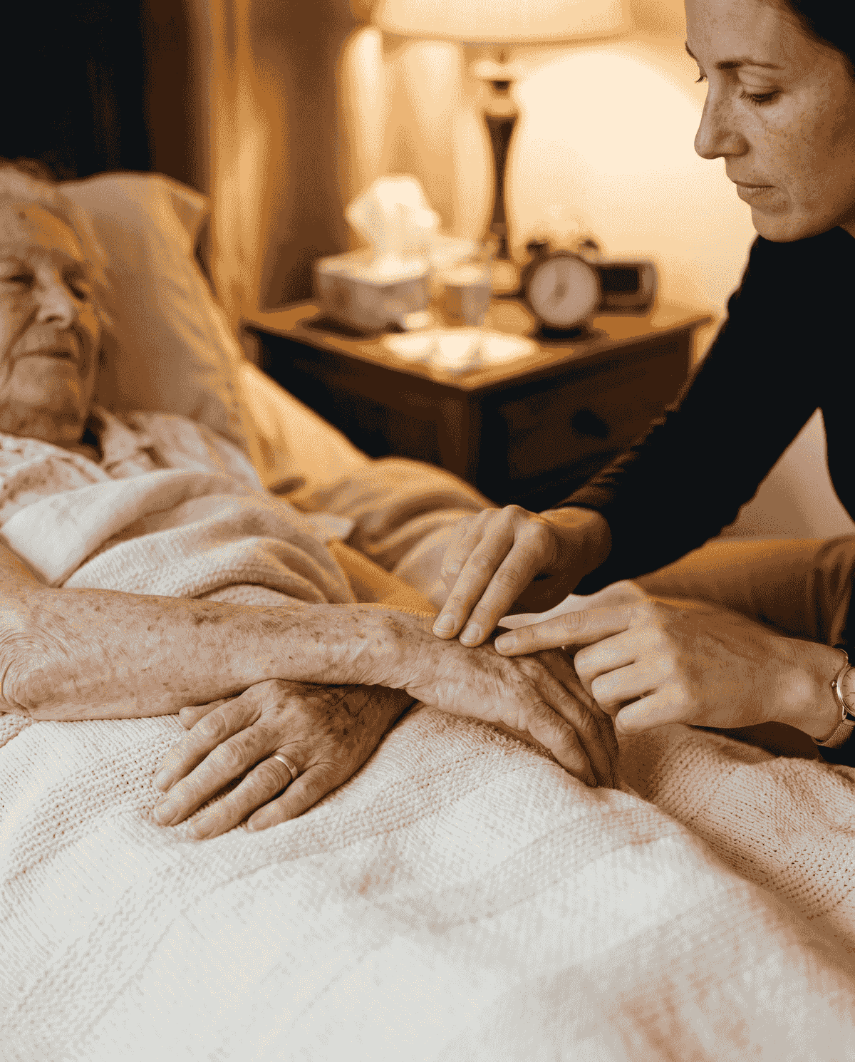 A caregiver at a softly lit bedside gently holding the hand of an older woman resting in bed, attentive to how she is doing