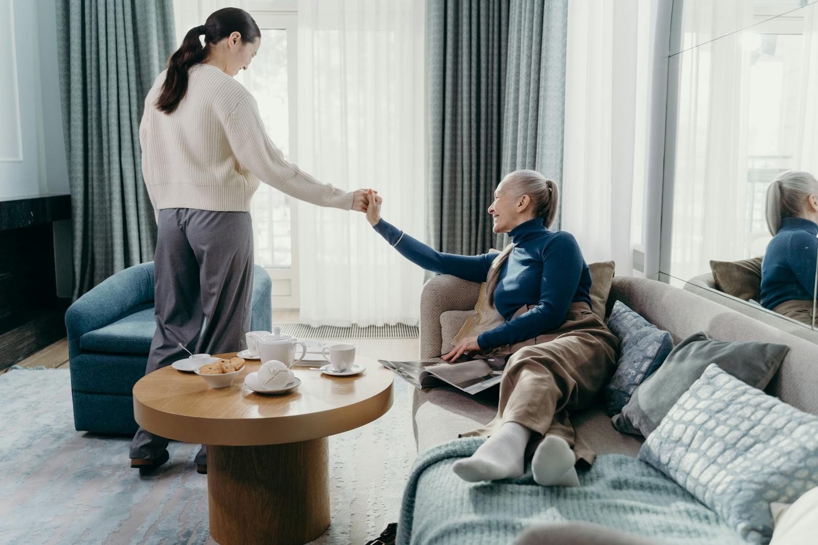 Adult daughter helping her elderly mother stand up from a sofa in a sunlit living room with tea and pastries on the coffee table, illustrating the daily physical work of family caregiving