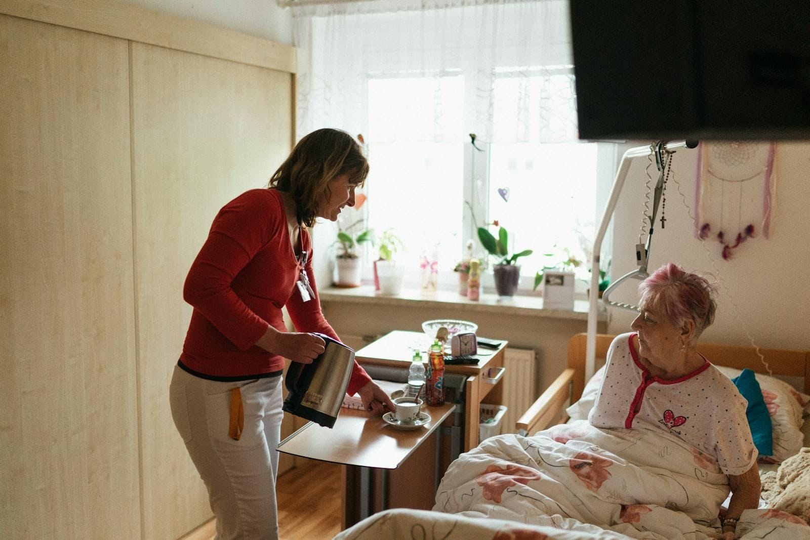 A trained caregiver in a warm sweater bringing a cup of tea to an elderly woman resting in bed in a sunlit bedroom decorated with potted plants and family touches