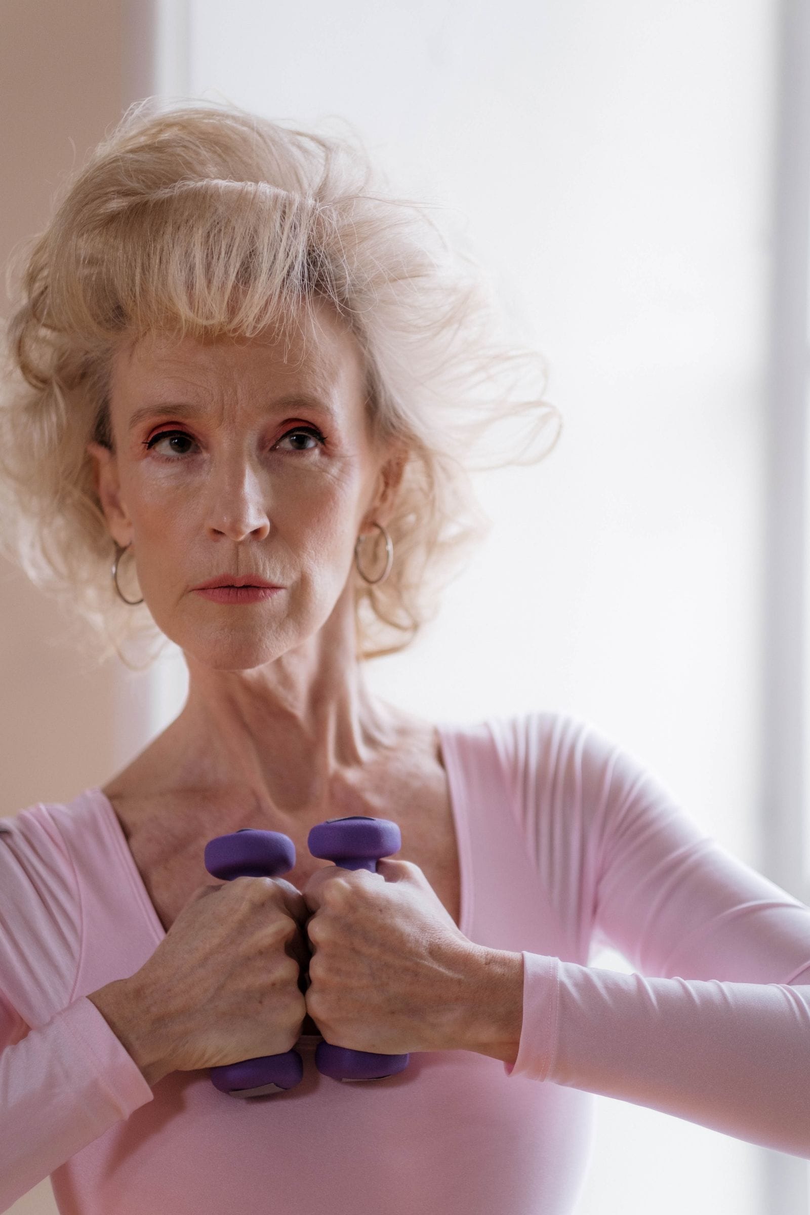 A focused white-haired older woman in a pink top holding a pair of small purple dumbbells at her chest