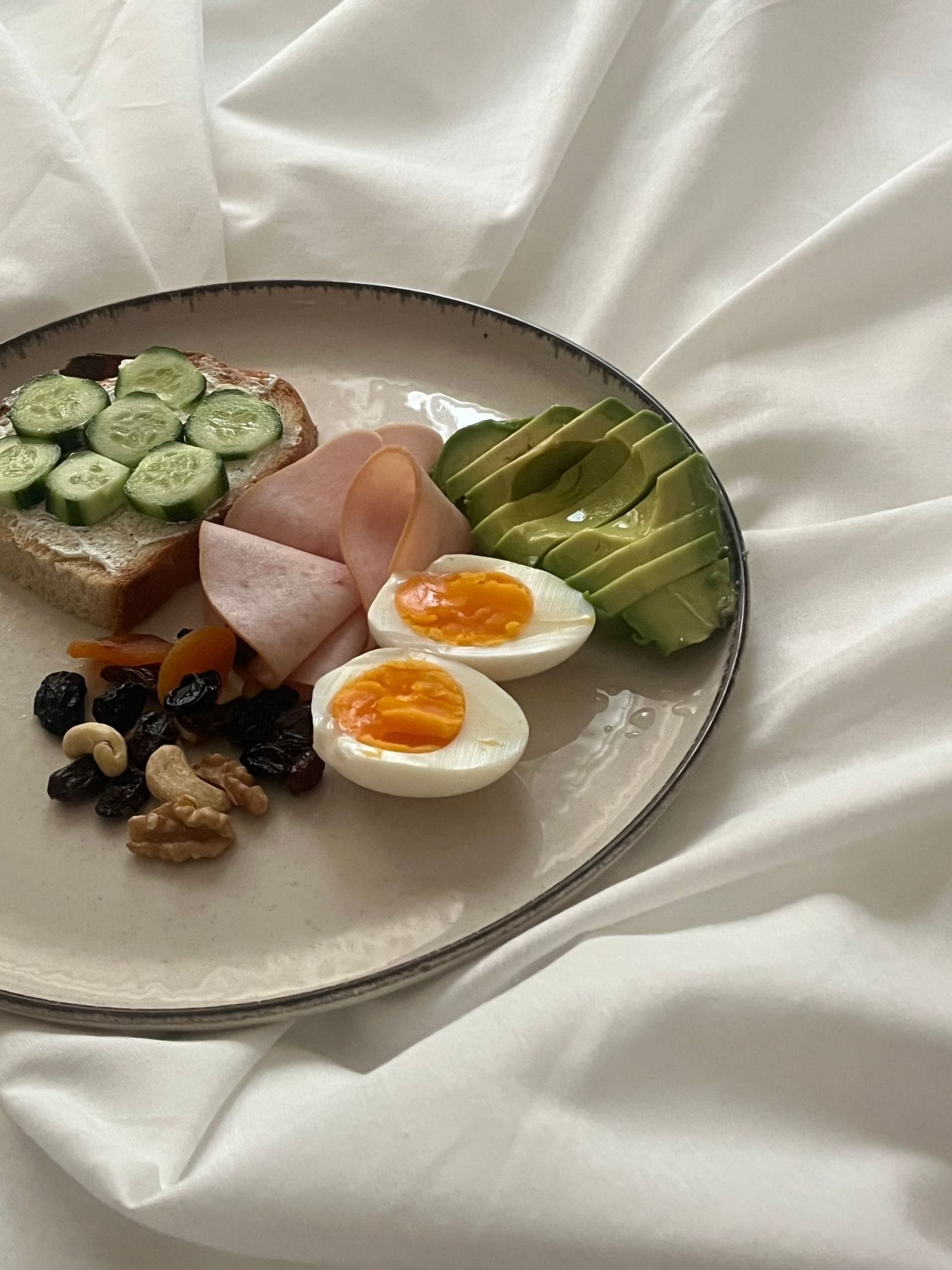 A plate of protein-rich foods including halved boiled eggs, sliced ham, avocado, mixed nuts, and dried fruit