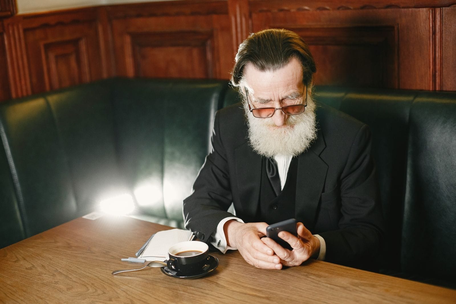 An older man in glasses sits at a wooden table reading a message on his phone with a cautious expression