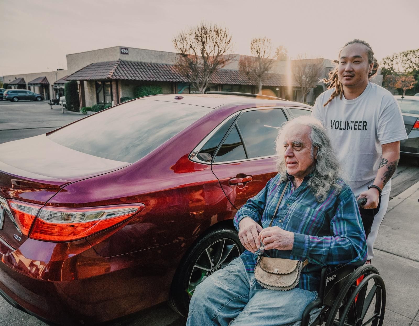 A volunteer driver in a blue T-shirt and jeans pauses next to an elderly man seated in a wheelchair near a row of parked cars on a sunny residential street, the helper's hands resting steadily on the chair's handles, both figures turned slightly toward the camera with quiet alertness