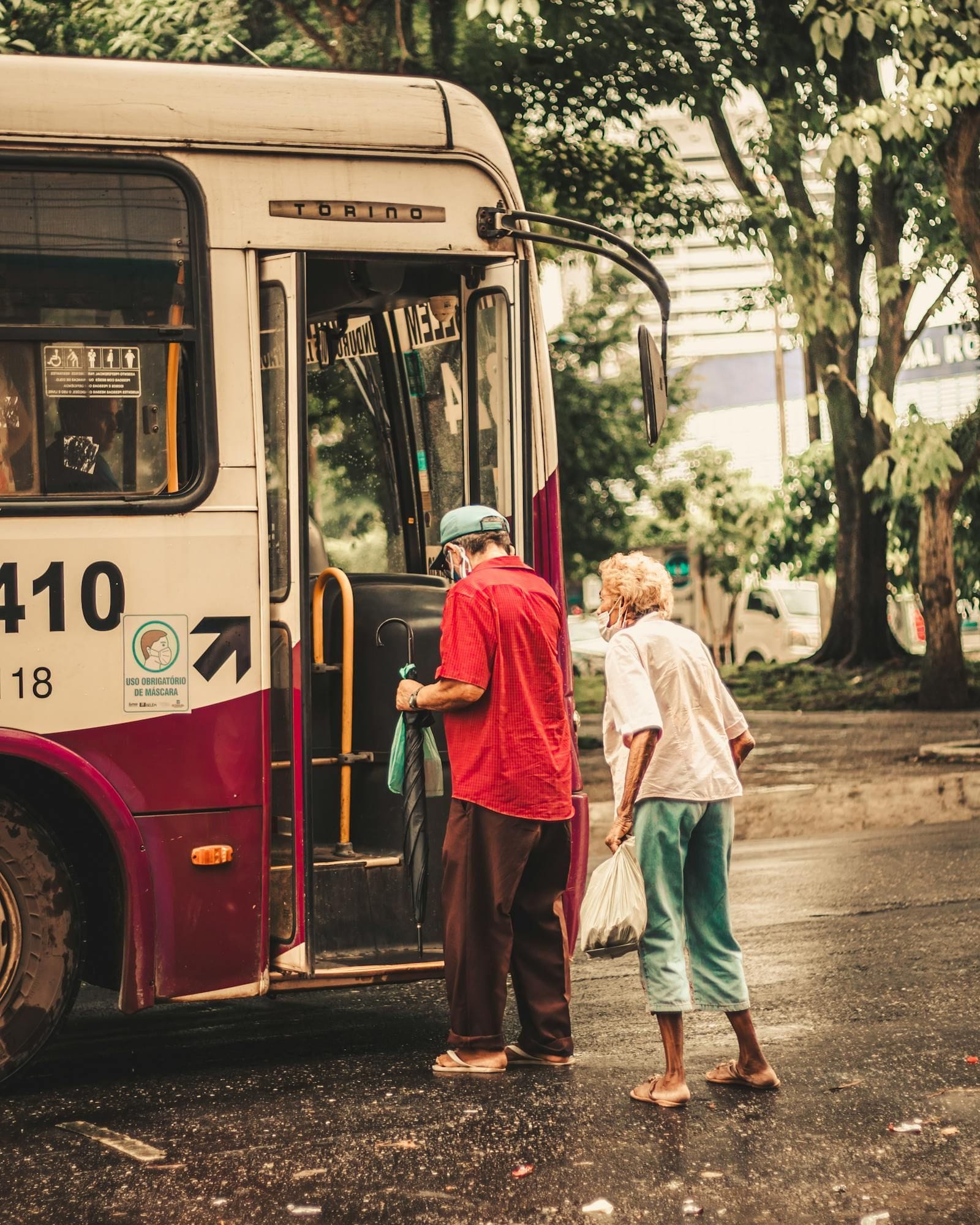 Two senior adults wait under a transparent shelter to board a city bus on a wet gray day, one in a dark coat with a small wheeled bag, the other in a long beige coat holding a closed umbrella, the bus's open accessibility door visible just past them