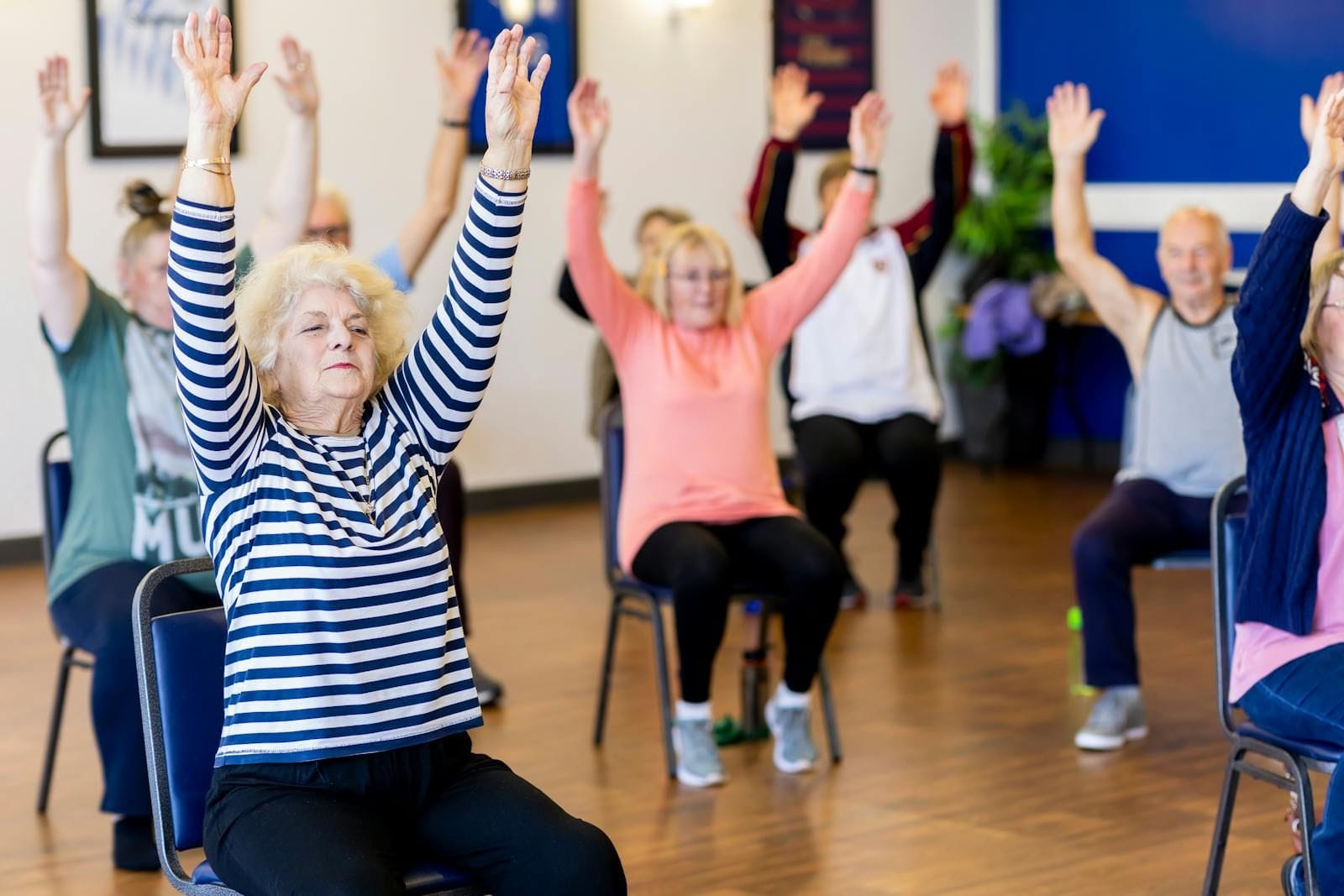 A group of older adults seated on chairs in a bright community-center class, arms raised overhead together during a gentle seated exercise session