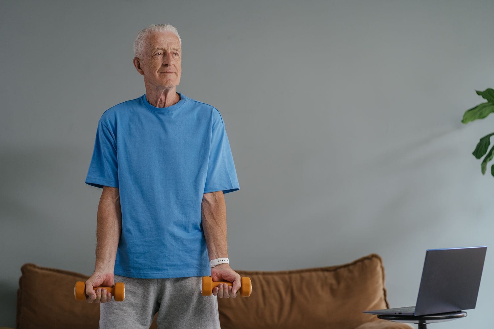 A white-haired older man in a blue shirt standing in his living room holding a pair of light orange dumbbells at his sides, a laptop on a stool beside him showing a workout to follow