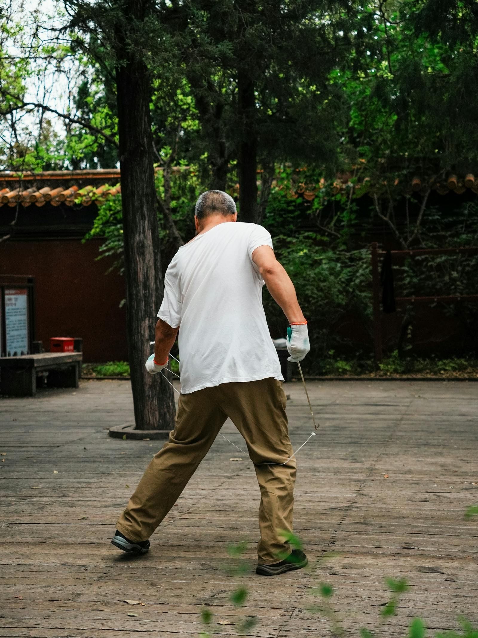 A silver-haired older man practices tai chi alone in a quiet park clearing, one arm extended at shoulder height and the other curved softly across his chest, his weight settled into a slightly bent rear knee as morning light filters through the trees behind him