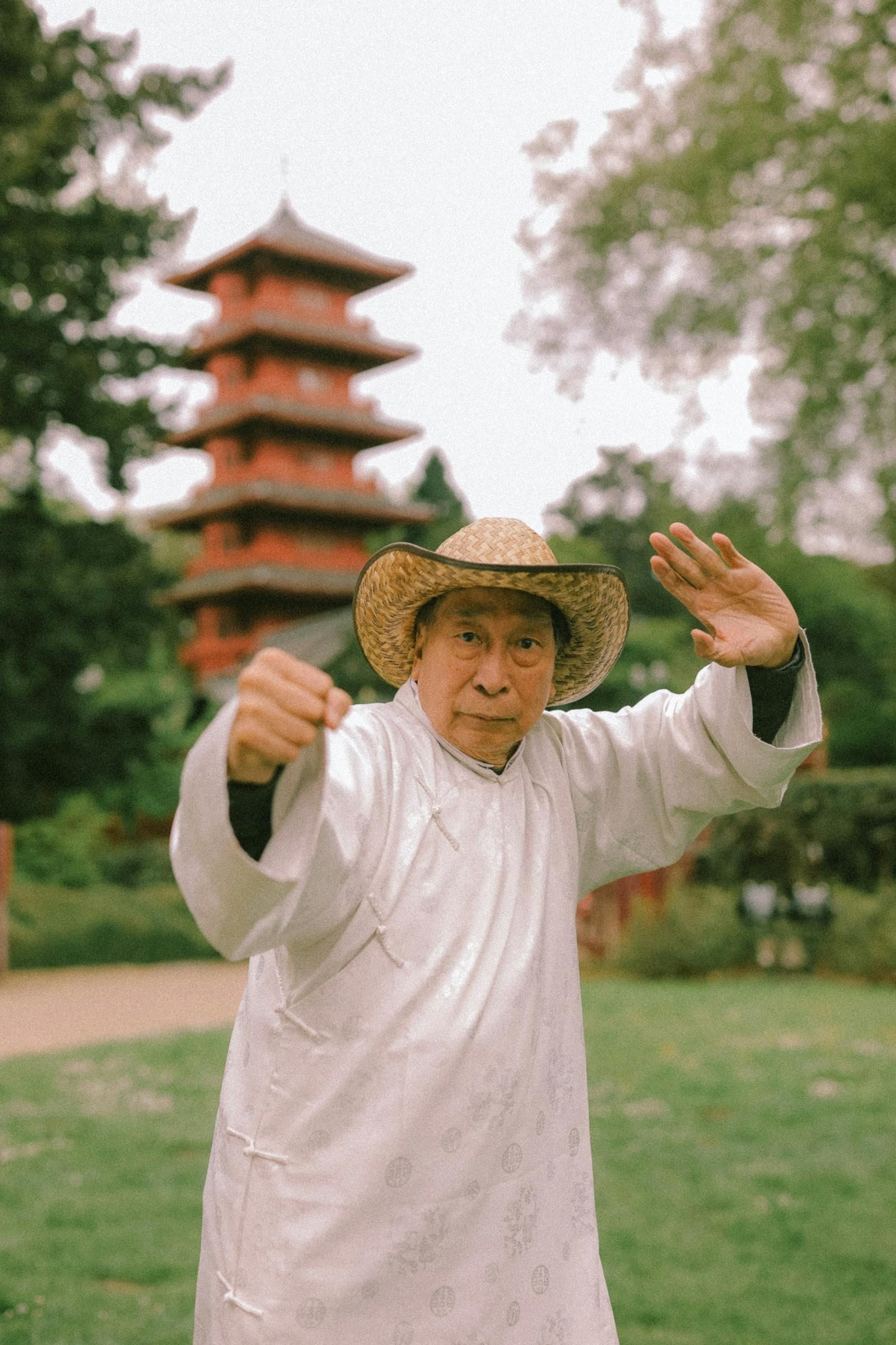 An older man with a white beard wearing flowing white traditional Chinese-style garments stands in a peaceful botanical garden, his right arm extended diagonally upward and his left hand held softly at his hip in mid-form, the deep greens of cultivated shrubs framing the quiet of the moment