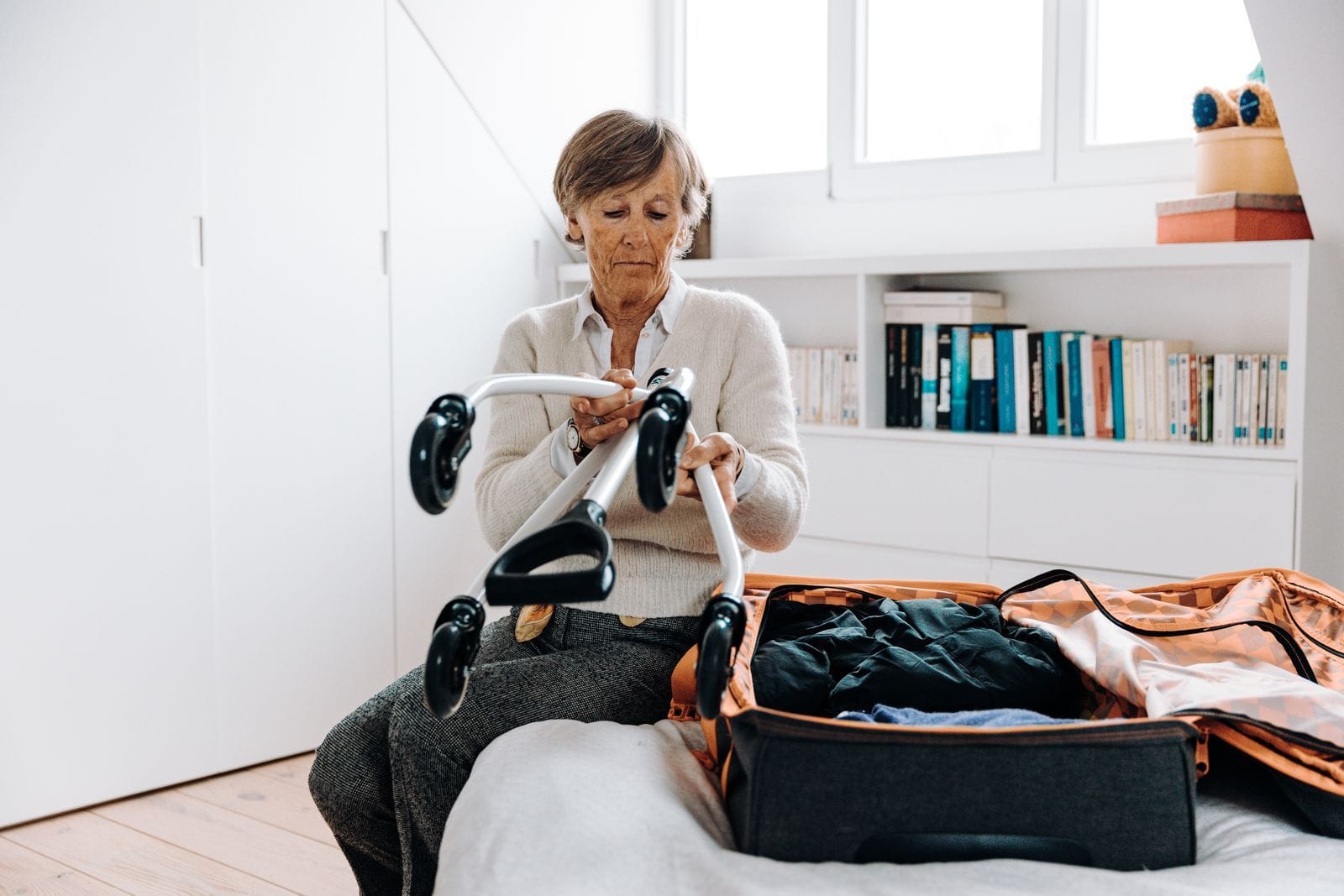 A silver-haired woman packing a suitcase open on the bed, her wheeled walker beside her in a bright bedroom