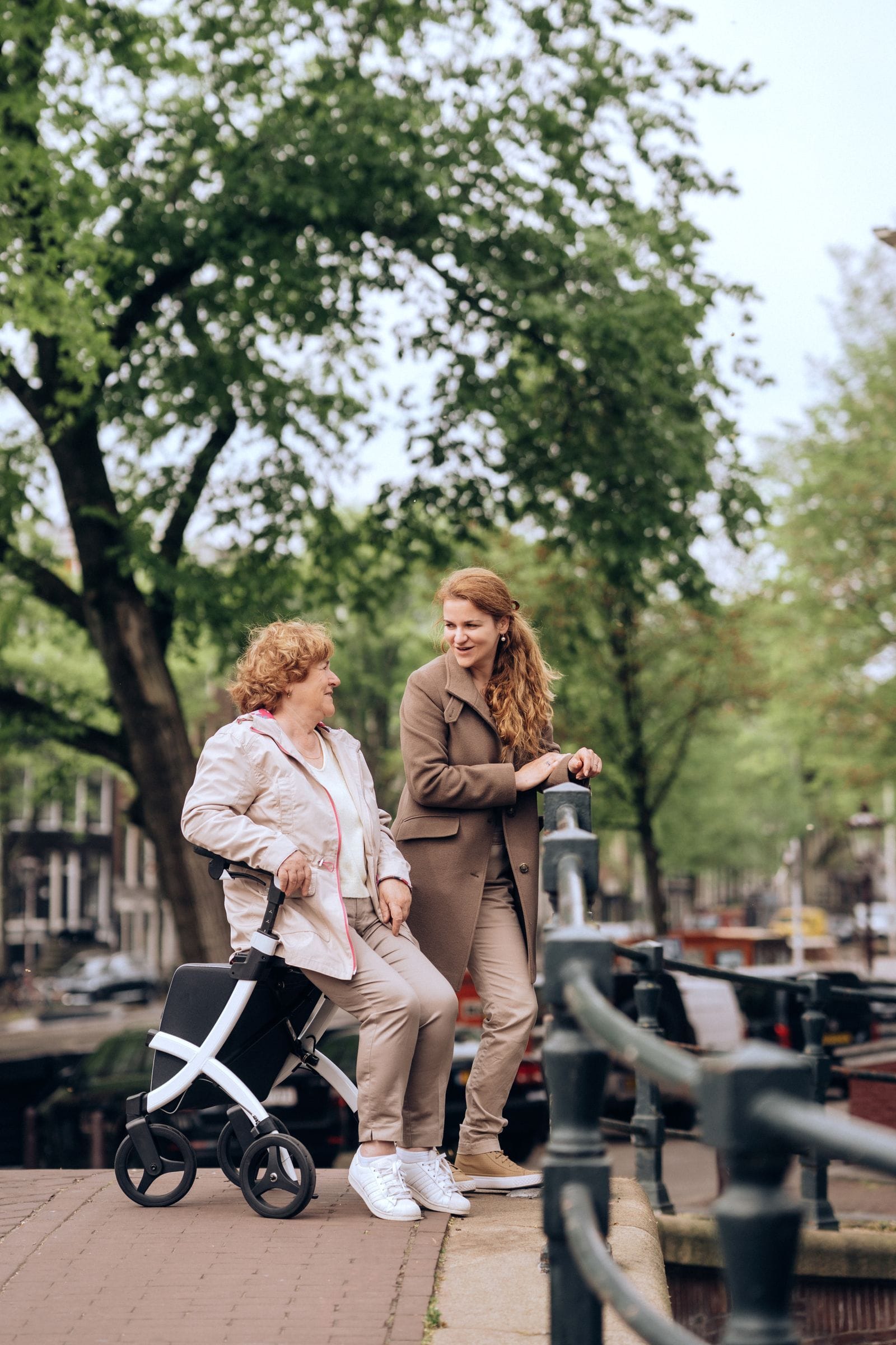 An older woman resting on her rolling walker outdoors, talking and smiling with her adult daughter on a bridge in soft daylight