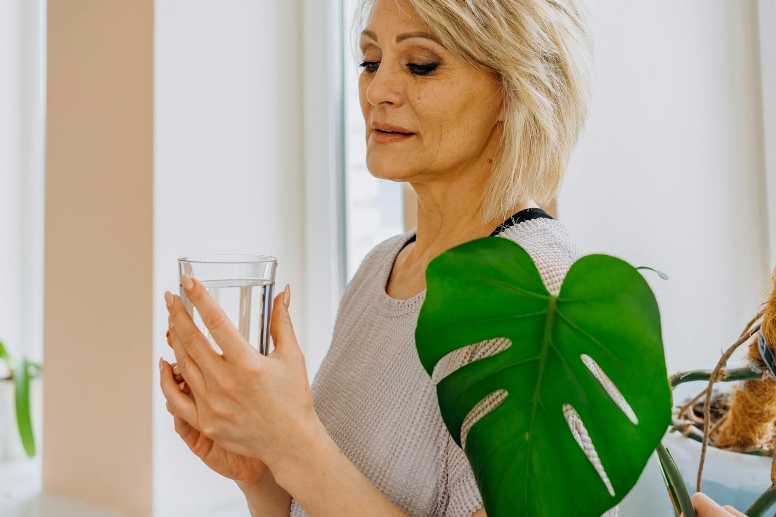 A senior woman with short blonde hair holds a clear glass of water near a bright window beside a green monstera plant, illustrating the role of hydration in preventing urinary tract infections in older adults