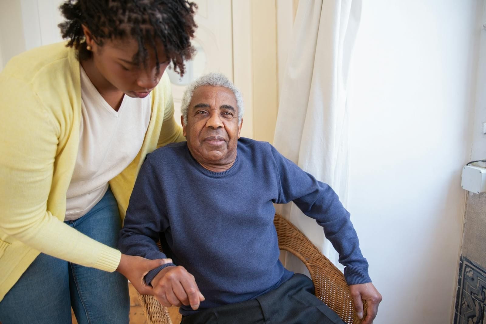 A kind caregiver in a yellow cardigan attentively assists an elderly man seated in a wicker chair near a sunlit window, illustrating the role of consistent in-home support in catching subtle health changes early