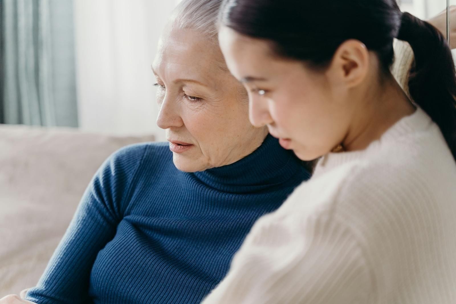 An adult daughter sits close beside her elderly mother in a softly lit indoor space, one hand resting gently on the older woman's, the two of them turned toward each other in the kind of unhurried conversation that families learn to have on the good afternoons