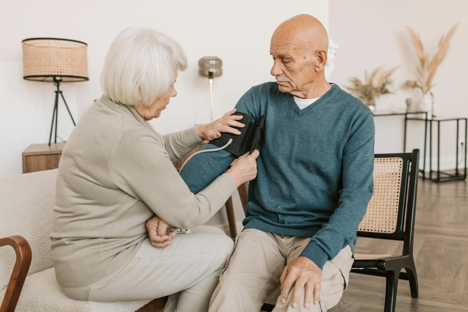 An elderly couple sits close together at a sunlit kitchen table, the wife wrapping the cuff of a small digital blood pressure monitor around her husband's upper arm with practiced care, the slow, ordinary ritual that becomes one of the most meaningful protections against the next step down