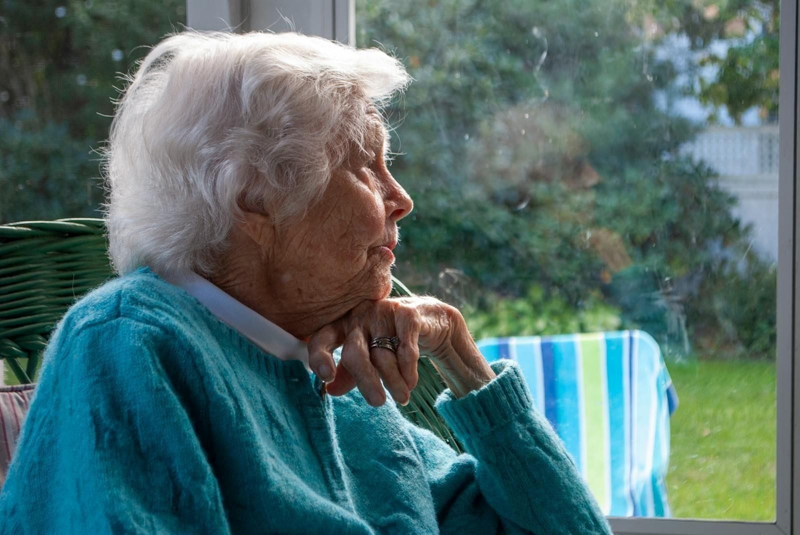 An elderly woman with white hair sits beside a sunlit window, her chin resting on her hand, gazing thoughtfully at something beyond the curtain, the kind of long, quiet stare that families of people with vascular dementia learn to recognize on the days when she is mostly here but not entirely