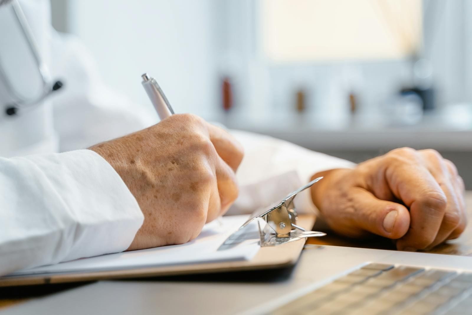 Close-up of an older professional in a white coat with rolled sleeves writing notes on a metal-clipped clipboard with a silver ballpoint pen, the freckled writing hand resting on the page in soft window light, the well-lit clinic background blurred behind them