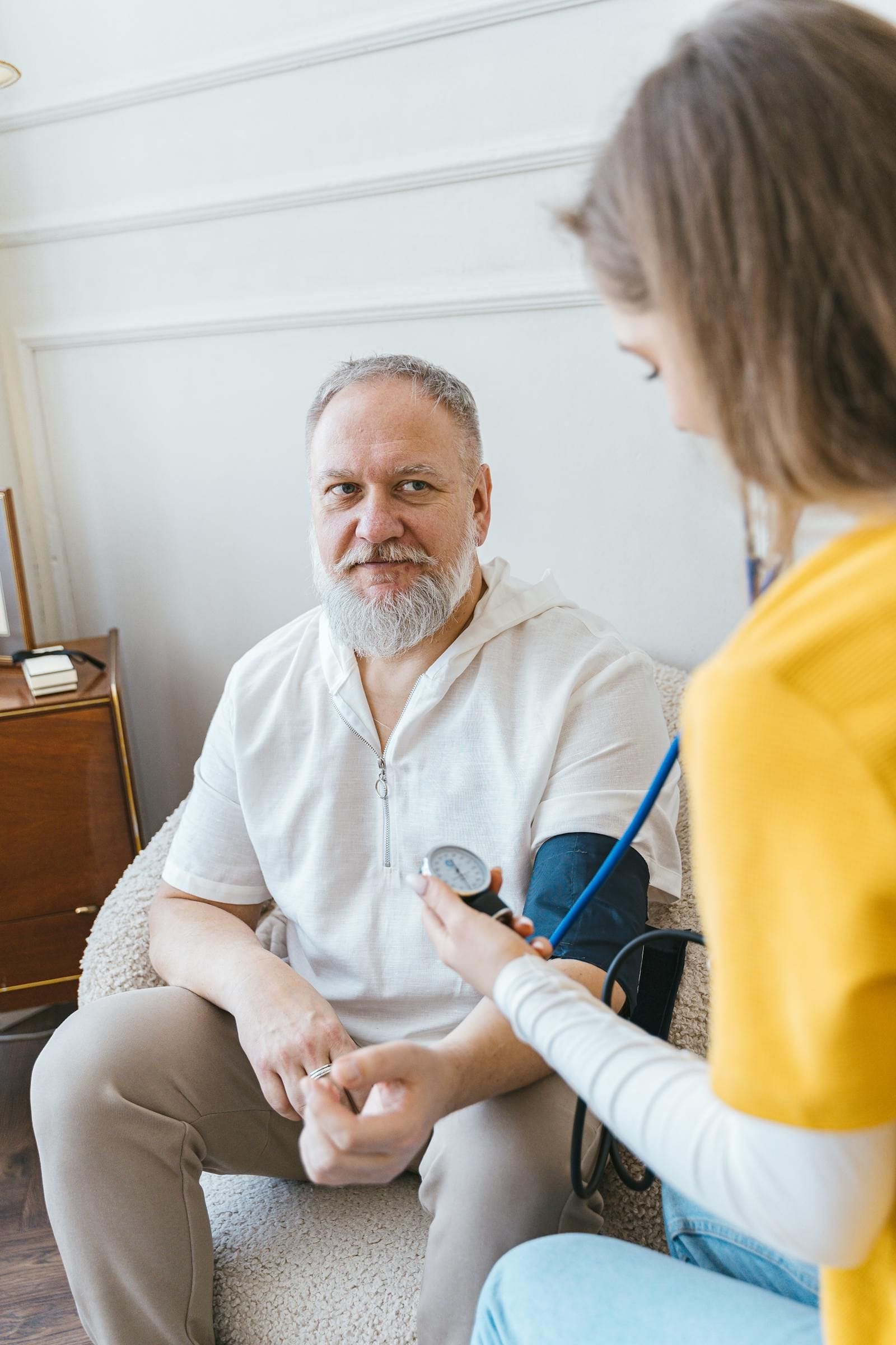 An older man with a closely-trimmed white beard and balding head in a white short-sleeve shirt sits in a soft upholstered armchair while a younger woman with brown hair and a mustard-yellow cardigan, her profile to the camera, takes his blood pressure with a blue-tubed stethoscope and a small black dial gauge attached to a cuff wrapped around his left arm