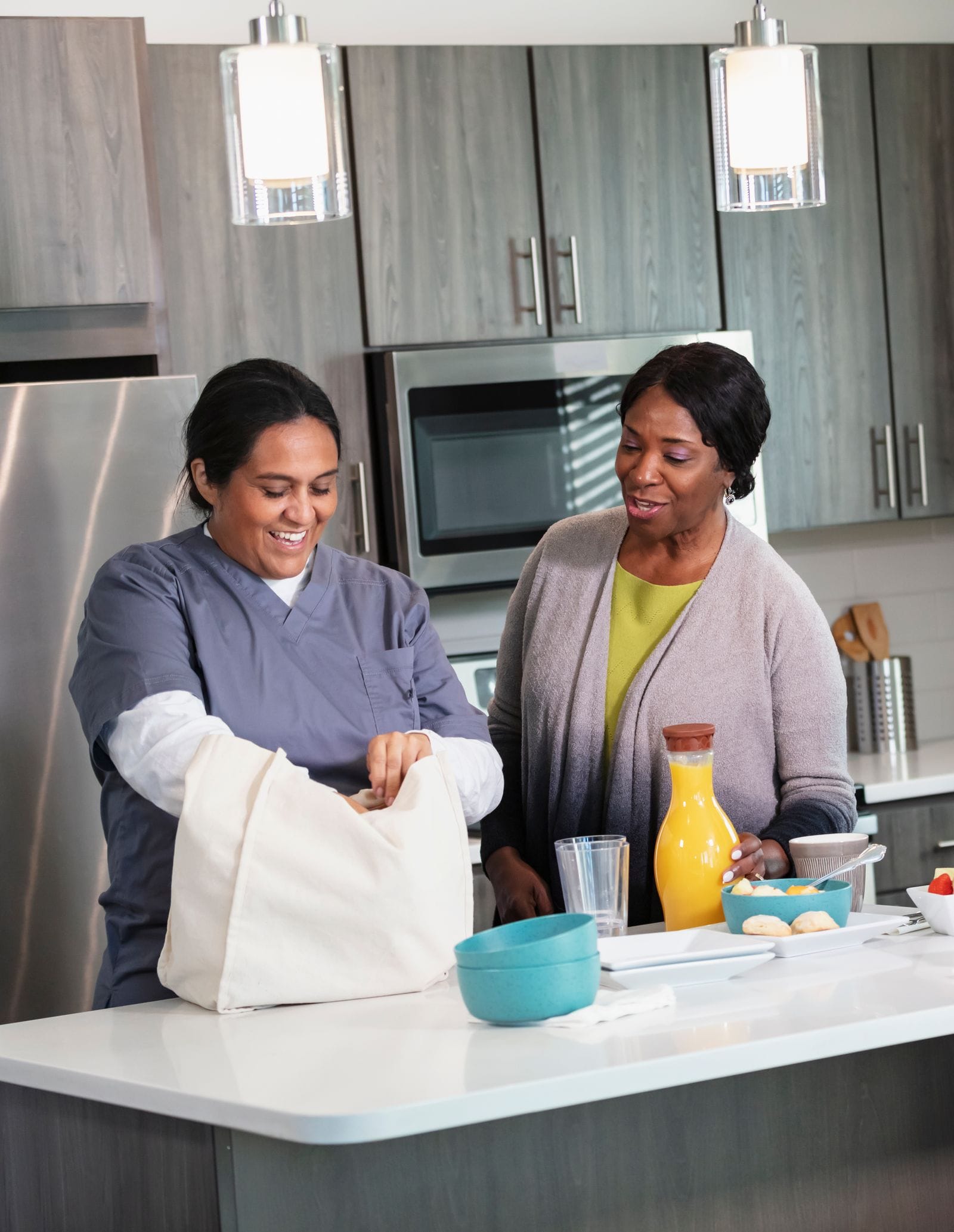 A home health aide unpacking groceries while chatting with an older woman in her kitchen